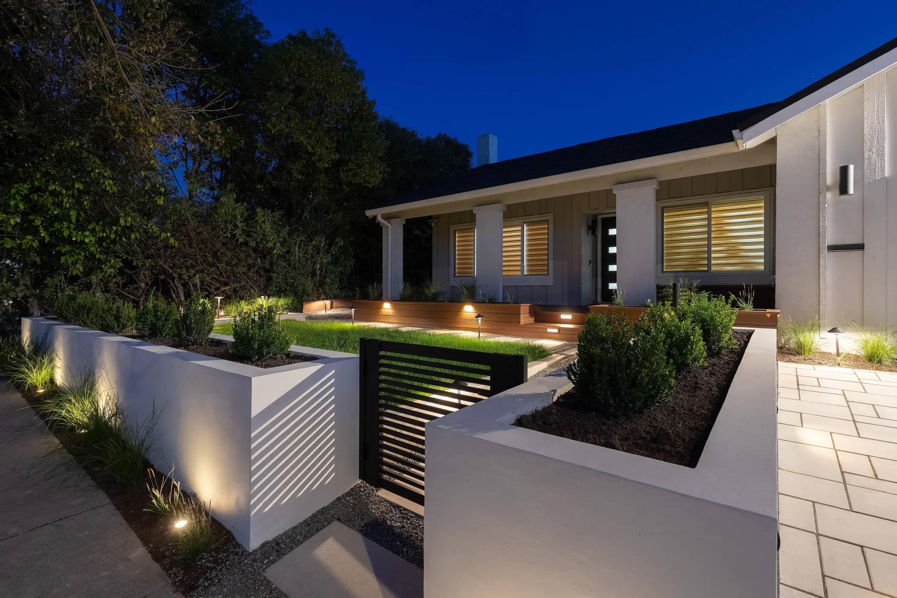 Modern home exterior with illuminated wooden deck, white planter walls, and landscaped entry at dusk