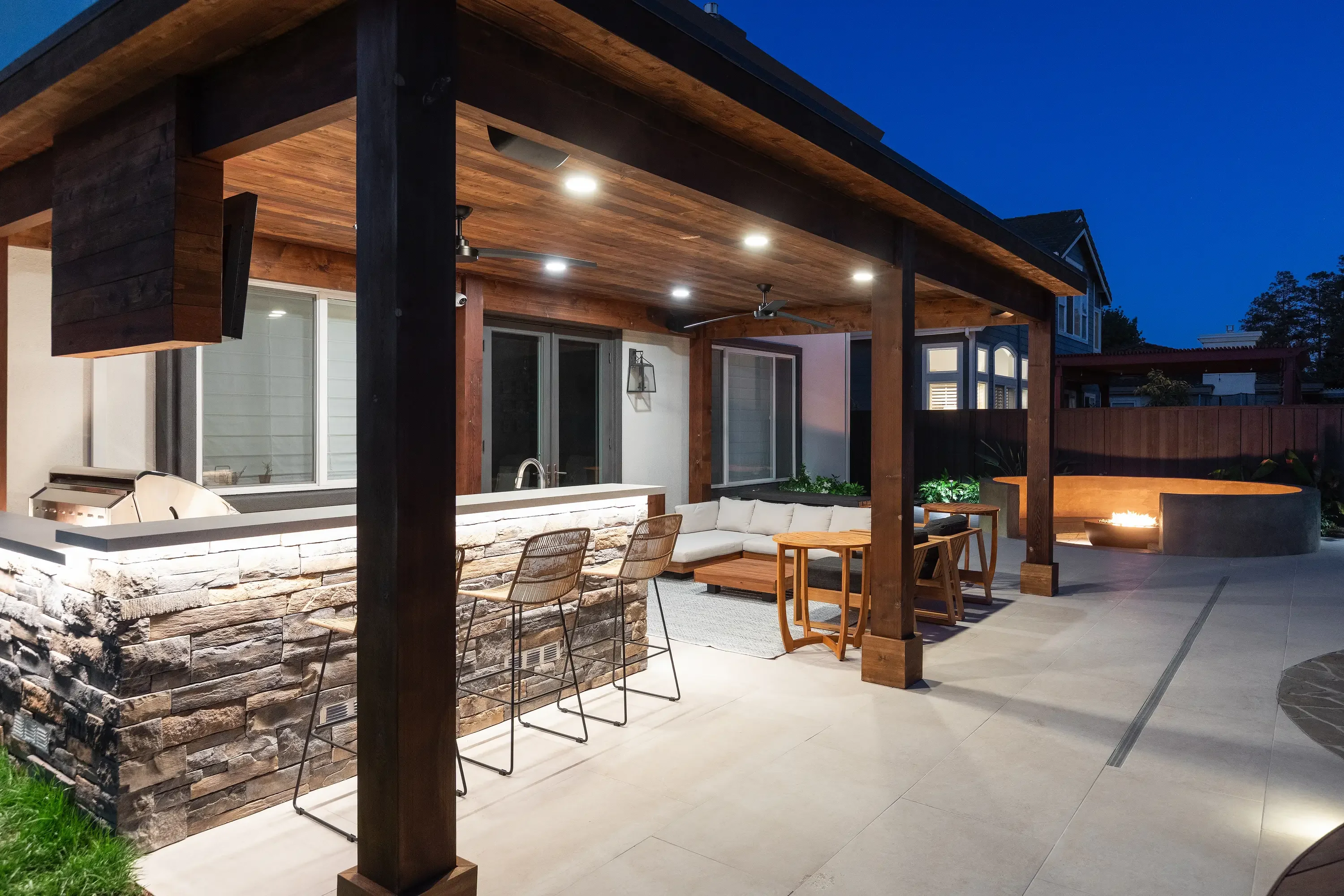 Wood pergola with LED lights, stacked stone kitchen, concrete patio, and fire pit at dusk