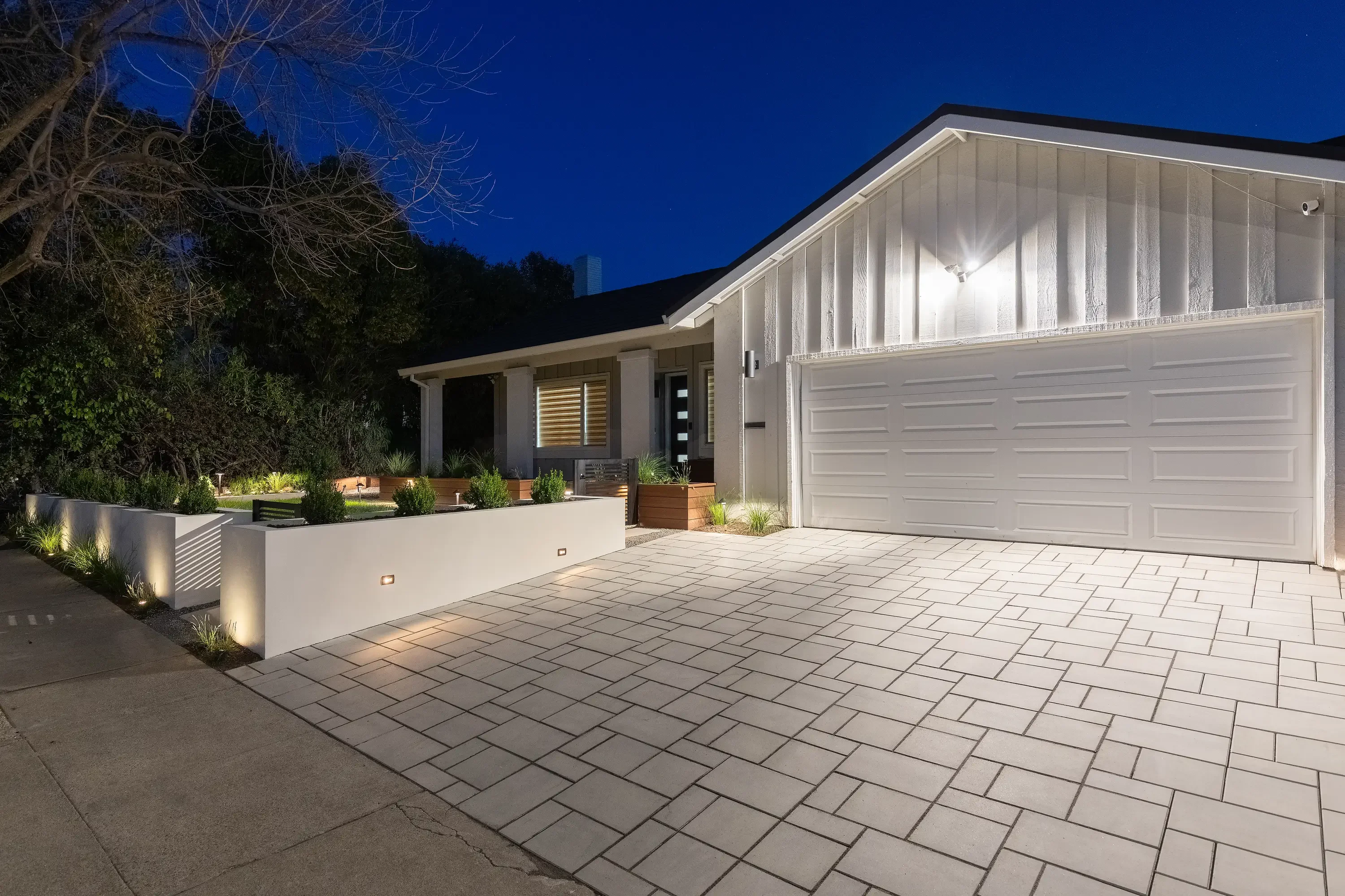 Modern home with newly installed paver driveway, raised planter walls, and landscape lighting at dusk