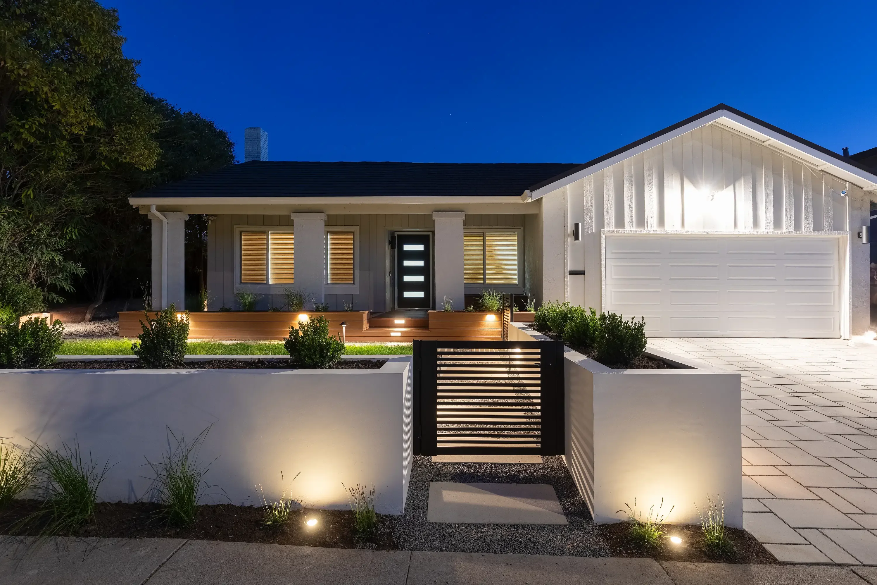 Modern home with illuminated white retaining walls, paved courtyard, and contemporary slatted gate at dusk