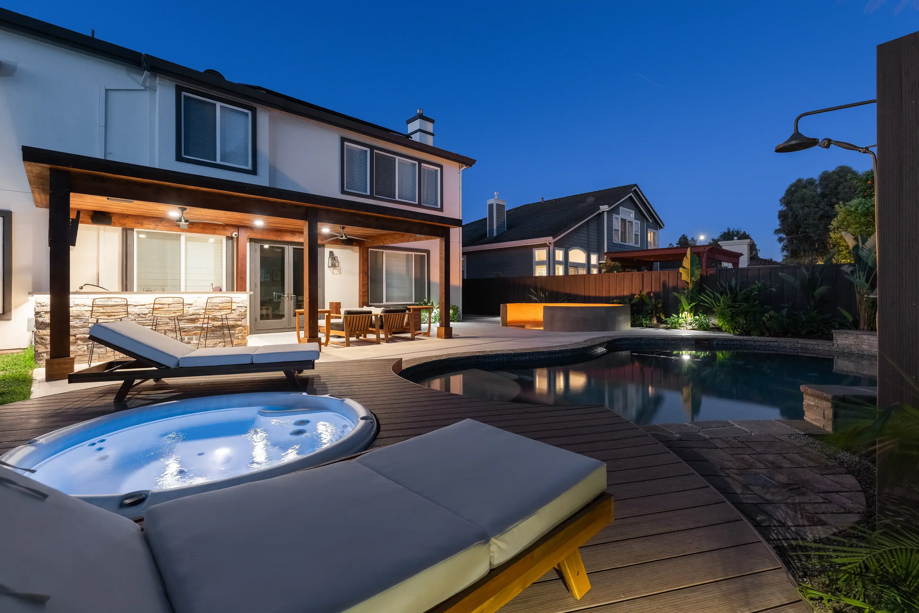 Composite deck with pool, spa, pergola, stone accents, and landscape lighting at dusk